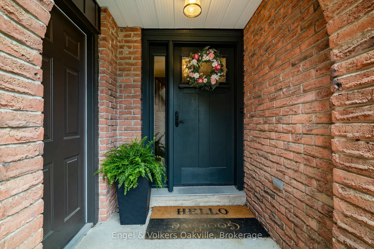 Brick home entrance with a dark door, floral wreath, and "Hello" doormat. A fern sits in a black planter.