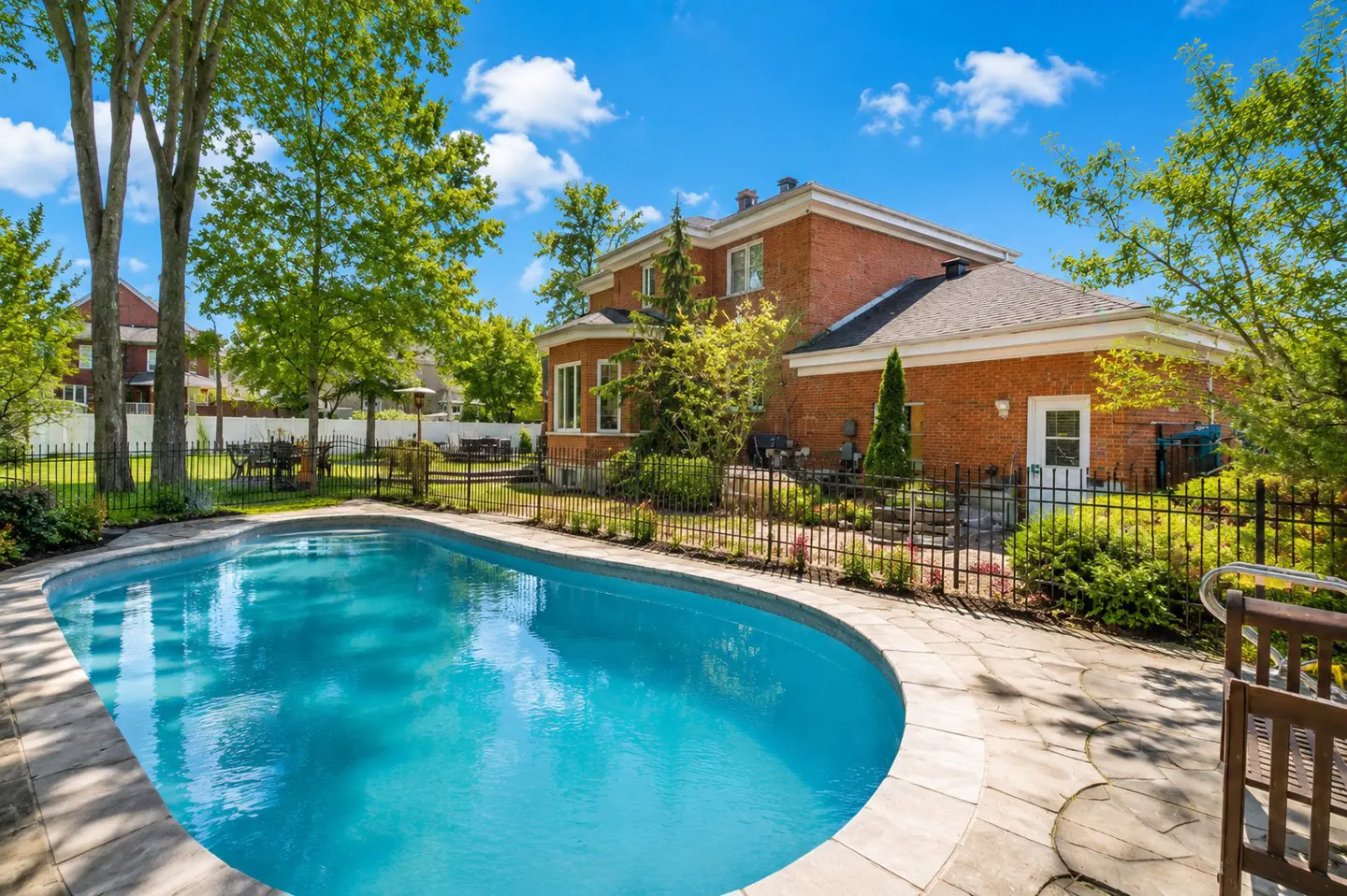Backyard view of a red brick house with a blue swimming pool, surrounded by green trees and a black metal fence.