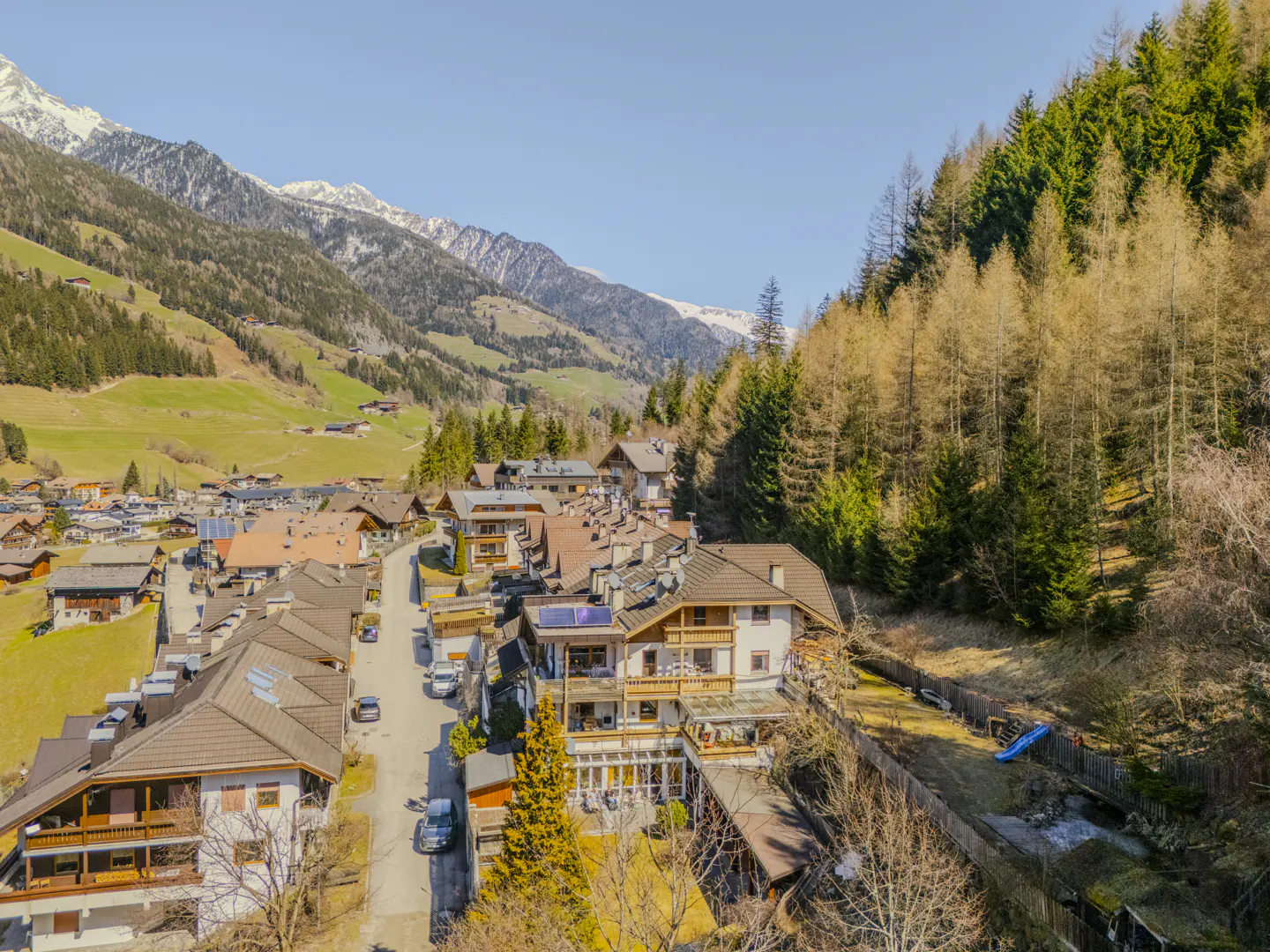 Aerial view of a village nestled in a valley, surrounded by mountains and forests under a clear blue sky.