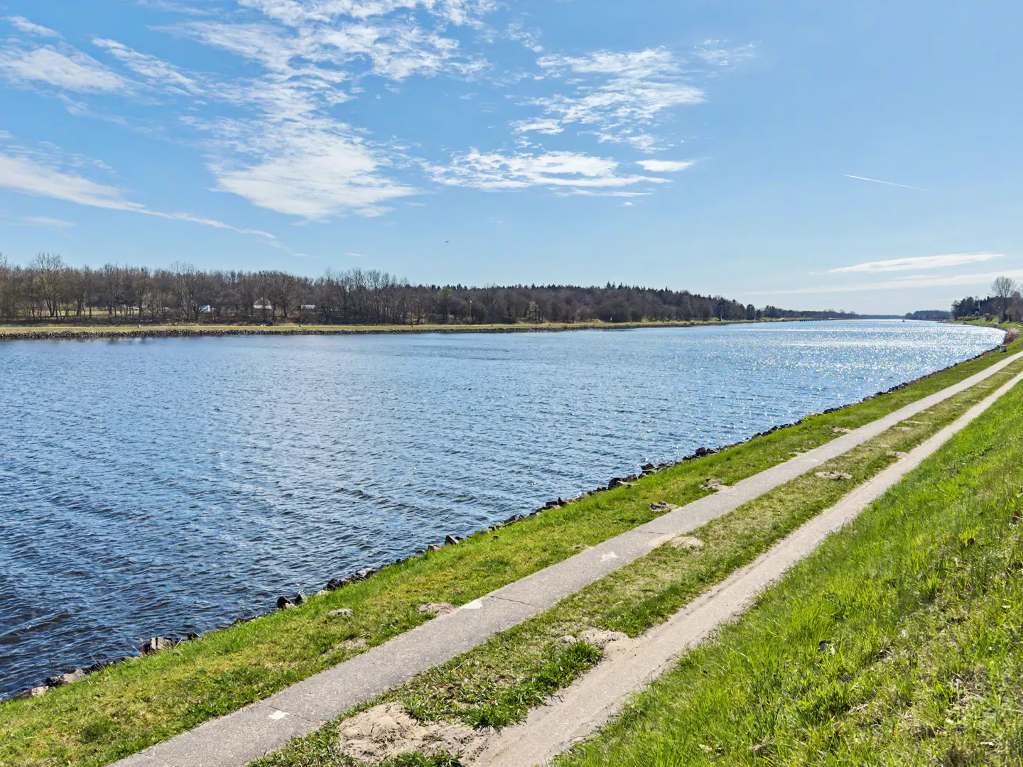 Scenic view of a wide blue river under a partly cloudy sky, with a grassy bank and a parallel walking path.