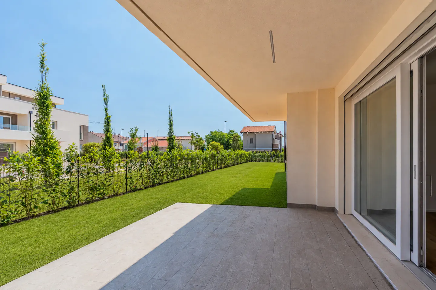 A modern home's patio with gray tiles leads to a green lawn and hedge. A white sliding glass door is on the right.