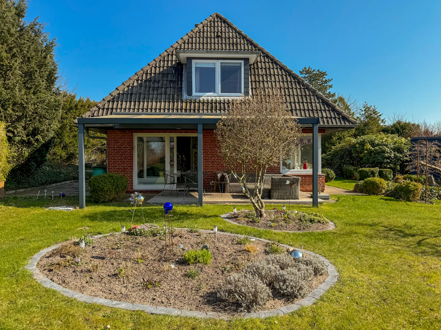 A red brick house with a gray tiled roof and a covered patio, surrounded by a green lawn and flower beds.