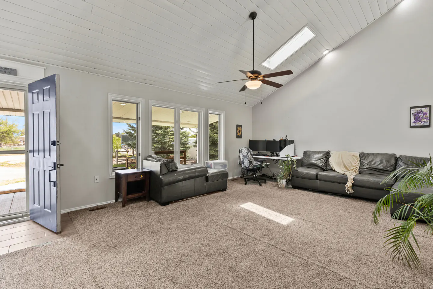 Living room with vaulted ceiling, gray sofas, and a desk with dual monitors. Open front door and large windows.