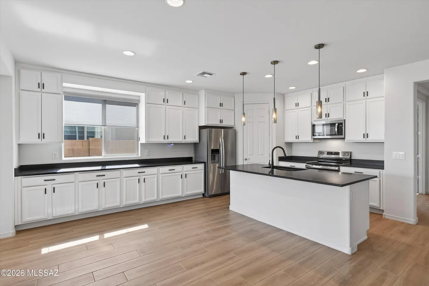 Bright kitchen with white cabinets, black countertops, stainless steel appliances, and wood-look tile flooring. Island with pendant lights.