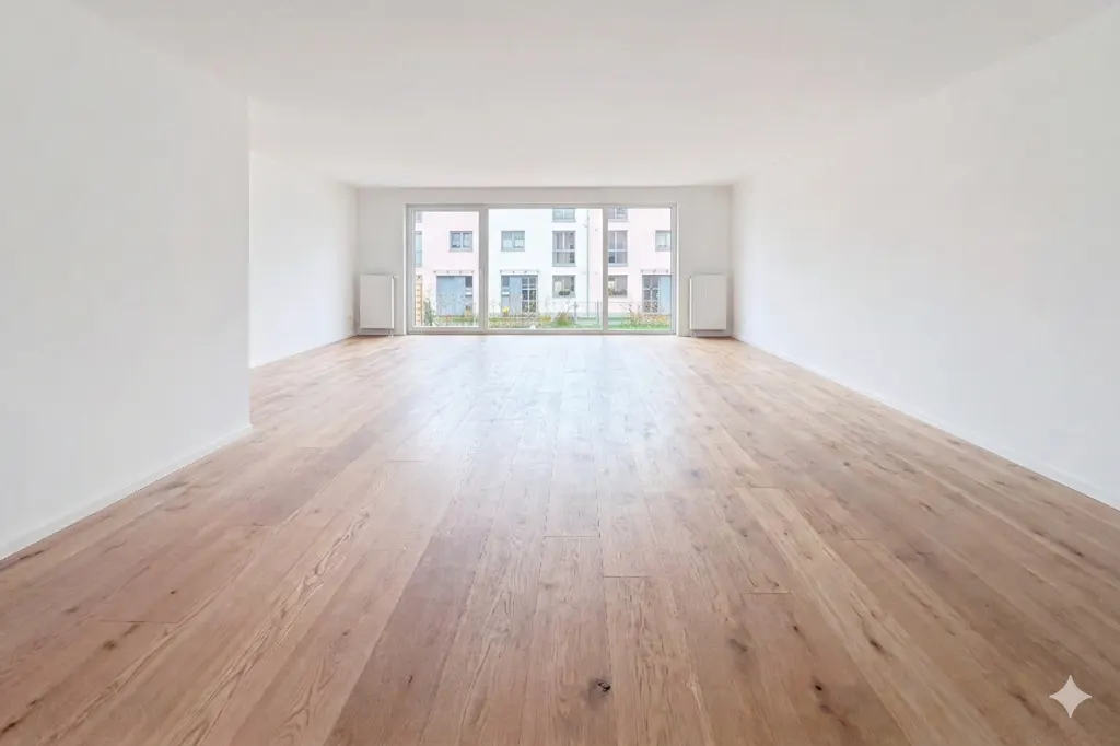 Bright, empty room with light wood floors and white walls. Large sliding glass doors offer a view of a building exterior.