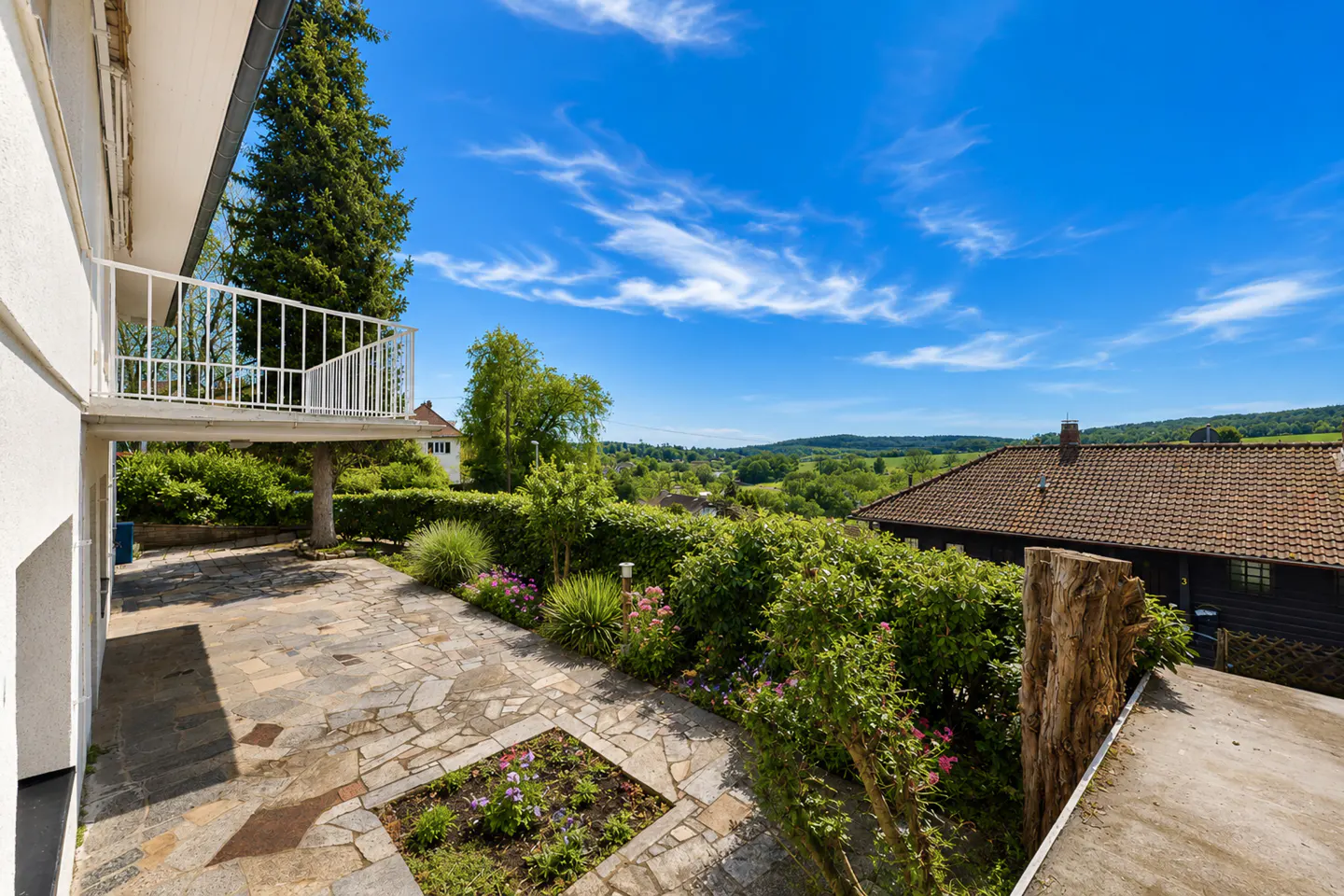 Stone patio with flower beds, hedges, and a white balcony under a blue sky. Rolling green hills in the distance.