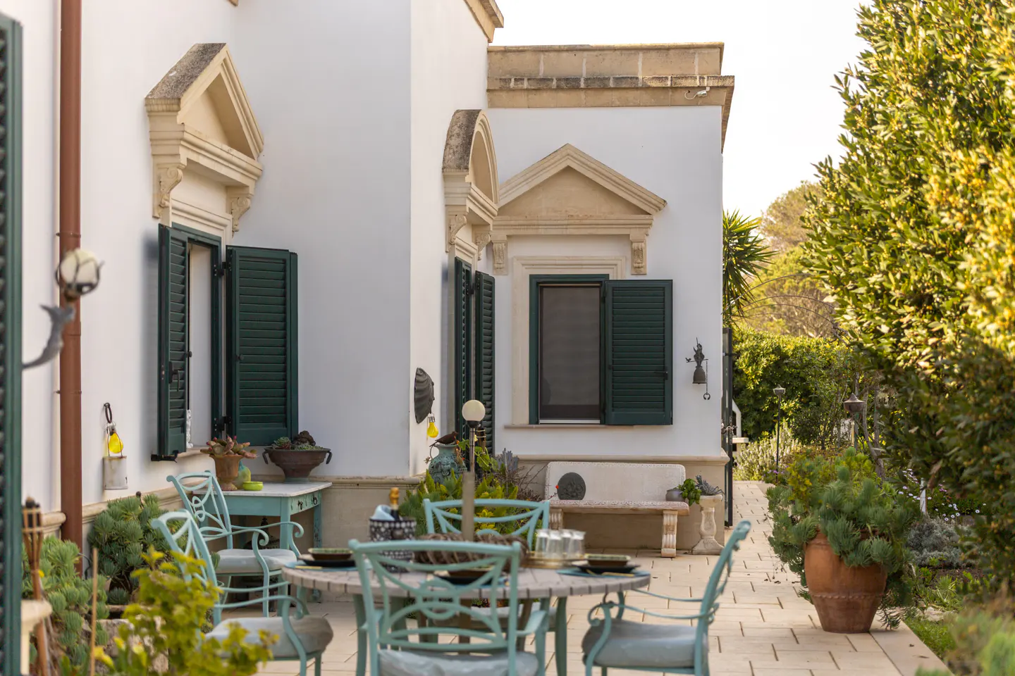 Outdoor patio with a round table, light blue chairs, and white building with green shutters in the background.