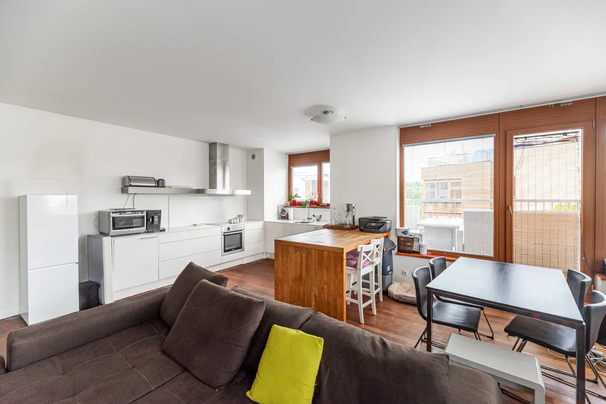 Open-concept apartment with white kitchen, wood island, brown sofa with yellow pillow, and black dining table near windows.
