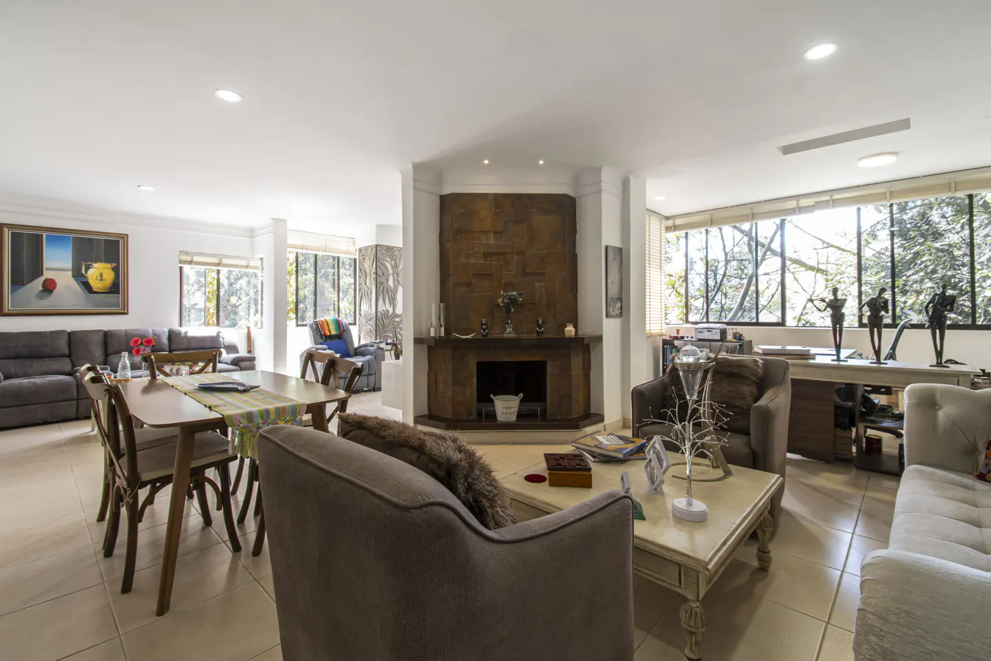 Bright, open living room with a stone fireplace, dining table, and large windows overlooking trees. Neutral colors and modern decor.