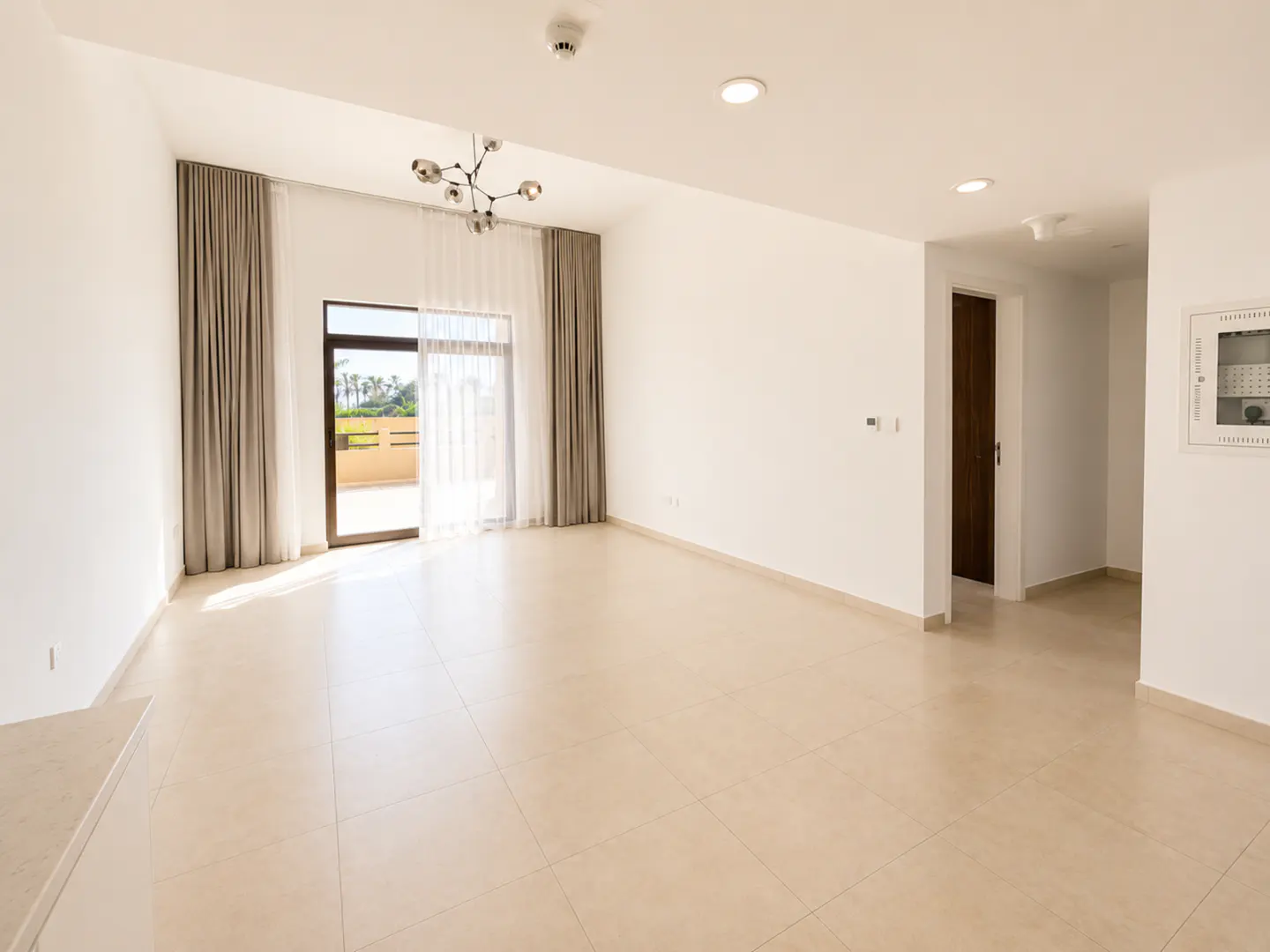 Bright, empty living room with beige tile floors, white walls, modern chandelier, and sliding doors to a balcony.