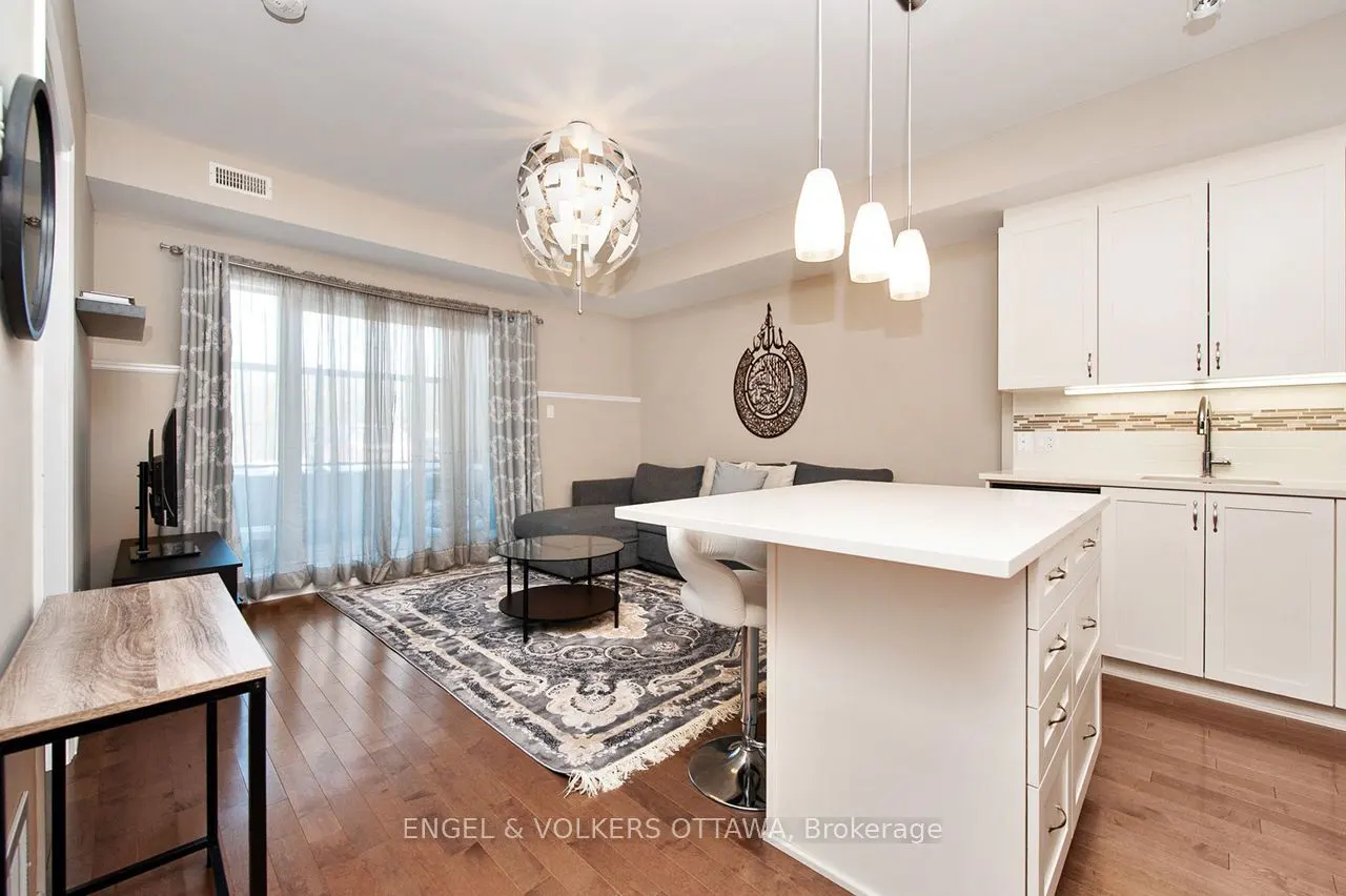 Bright condo interior with hardwood floors, a grey sectional sofa, and a white kitchen island with pendant lights. A patterned rug sits under a glass coffee table.