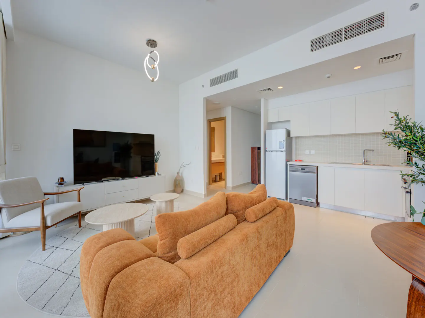 Bright, modern apartment with orange sofa, white walls, TV, and kitchen. A white chair sits on a patterned rug.