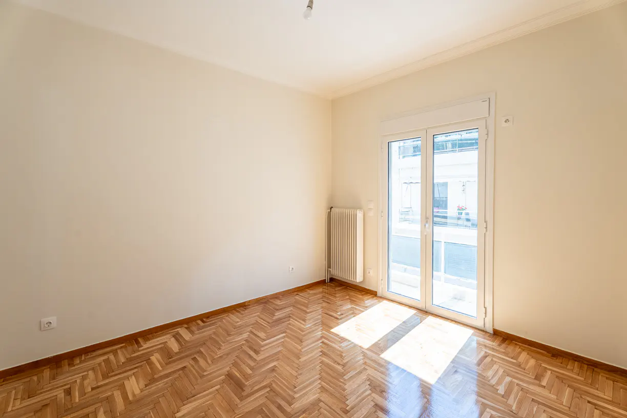 Empty room with herringbone wood floors, cream walls, and a white radiator. A glass door leads to a balcony with a city view.