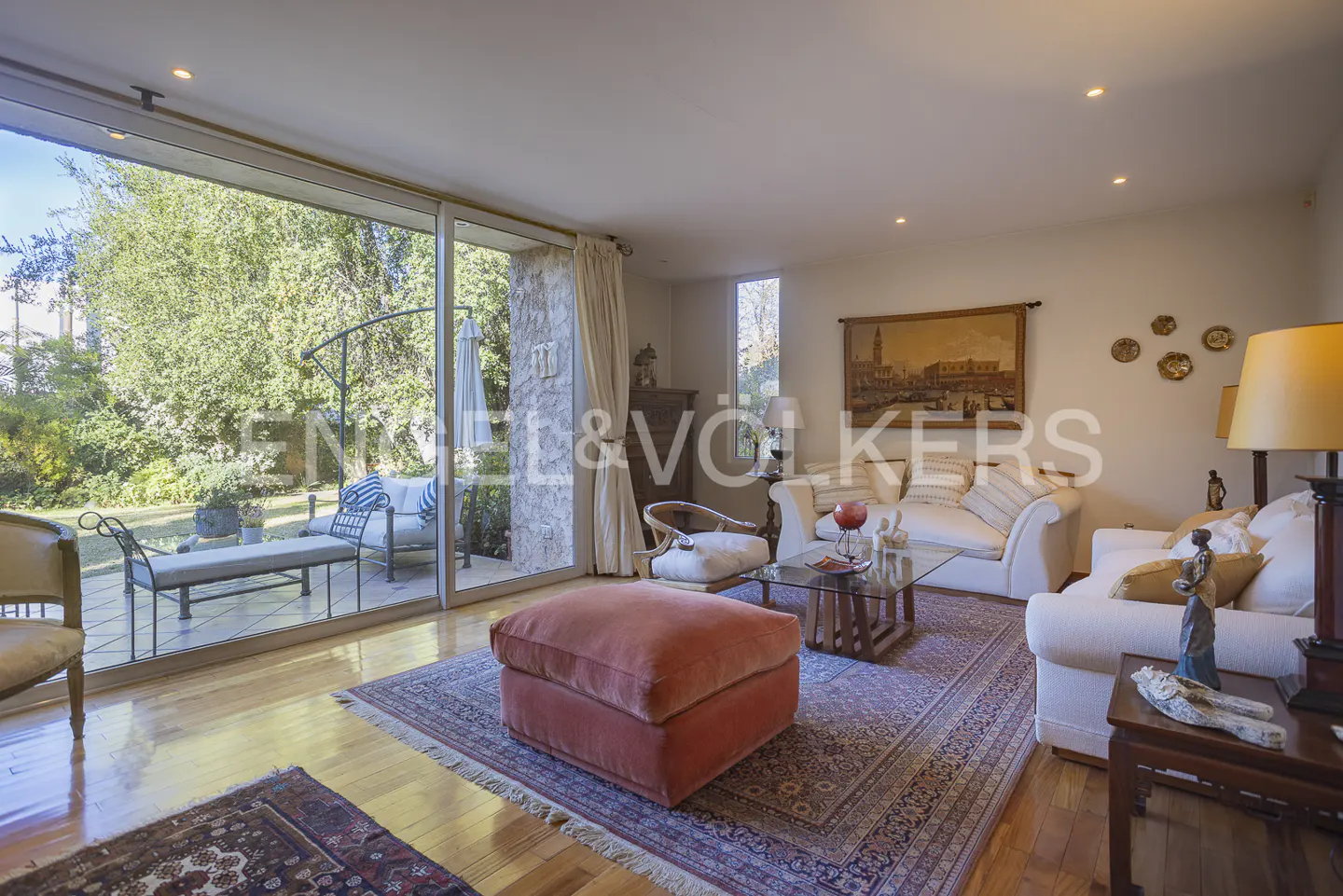 Bright living room with a large glass door, a coral ottoman, white sofas, and a patterned rug on hardwood floors.