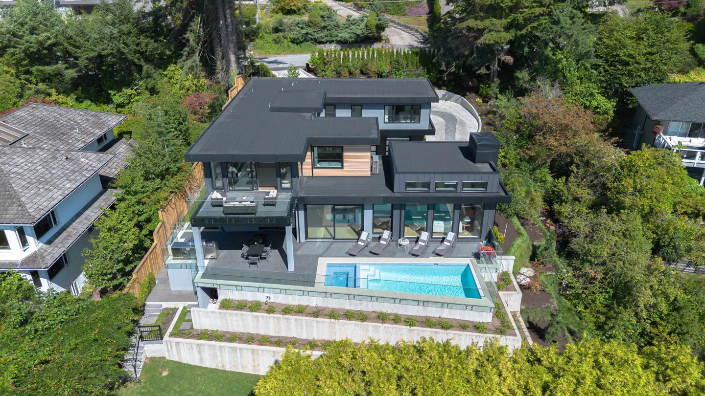 Aerial view of a modern, multi-story home with a dark roof, a pool, and a patio with lounge chairs. Lush greenery surrounds the property.