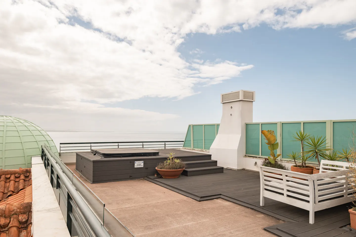 Rooftop terrace with a hot tub, white chimney, and potted plants. Ocean view and cloudy sky in the background.
