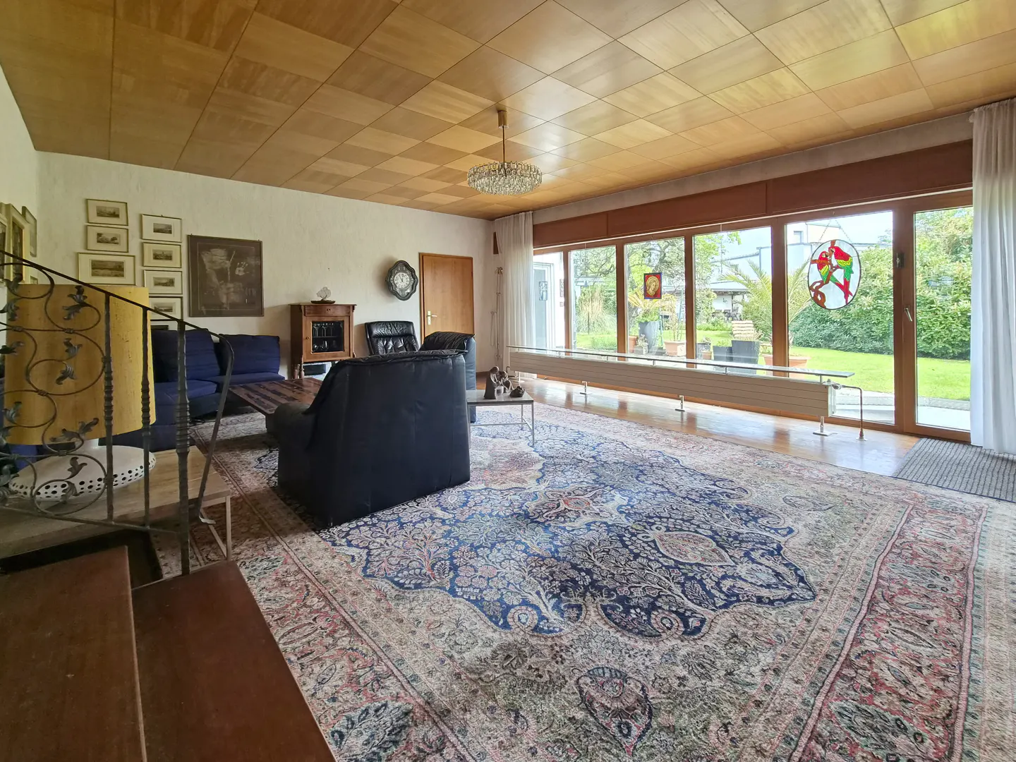 A living room with a large patterned rug, blue sofas, and a wall of windows overlooking a green lawn.