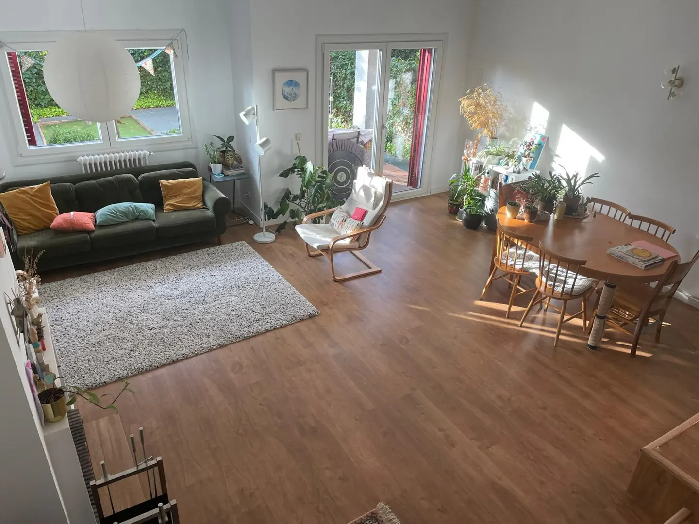 Bright living room with wood floors, a green sofa with colorful pillows, a white chair, and a wood dining table with chairs.