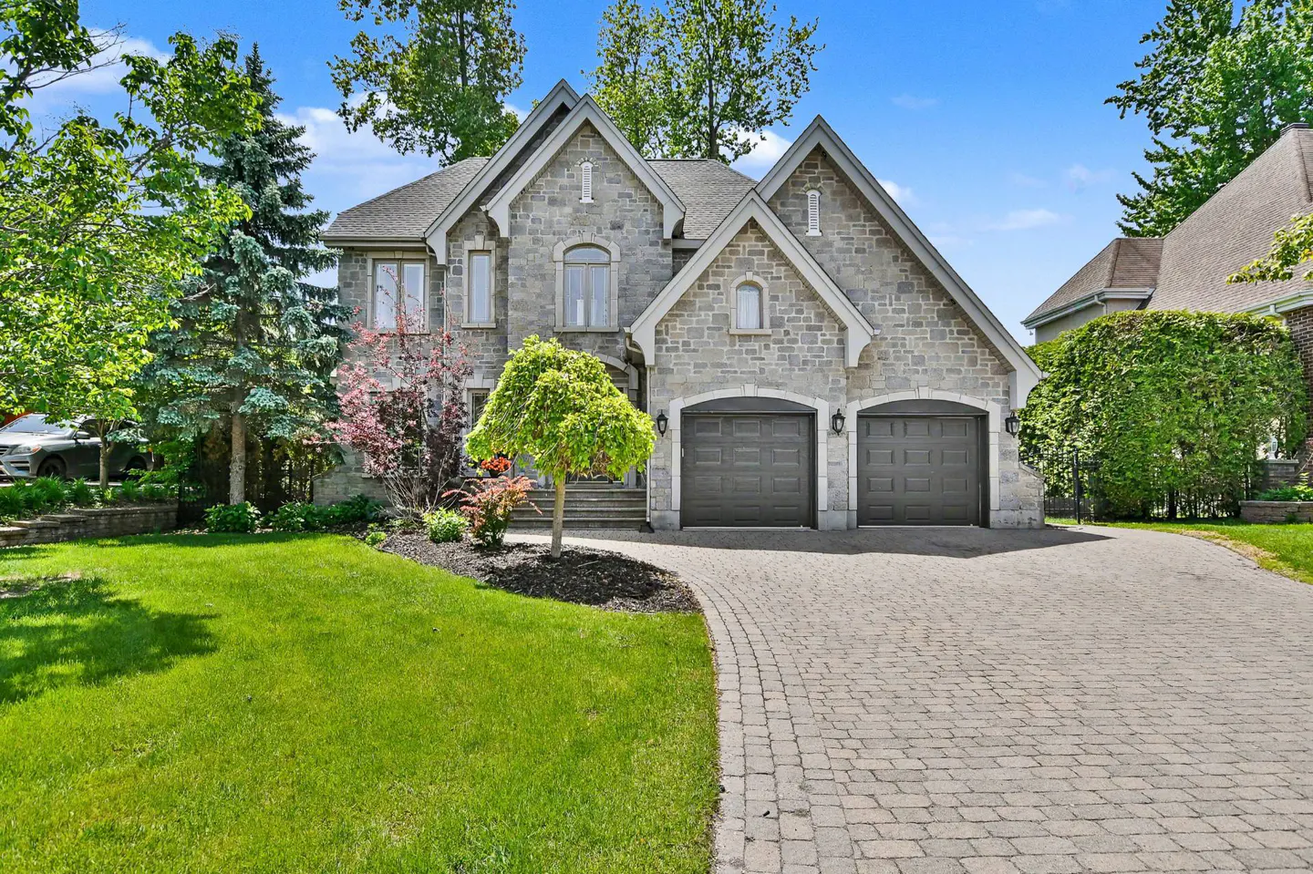 Two-story stone house with a double garage, paved driveway, and lush green front lawn under a clear blue sky.