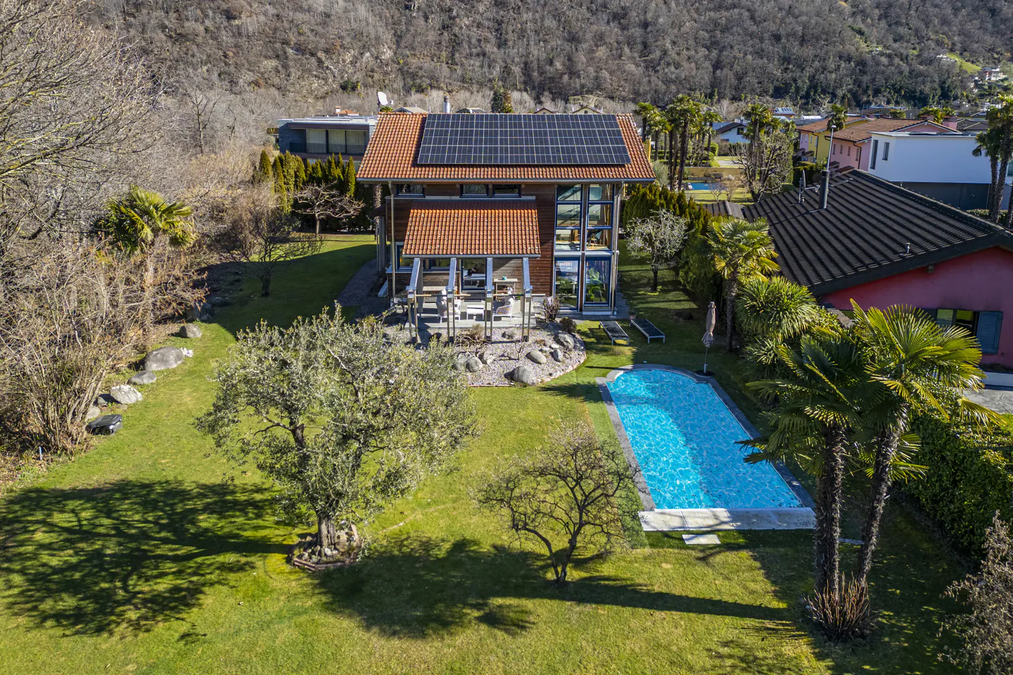 Aerial view of a modern house with solar panels, a pool, and a large green lawn. The house has a red tile roof and large windows.