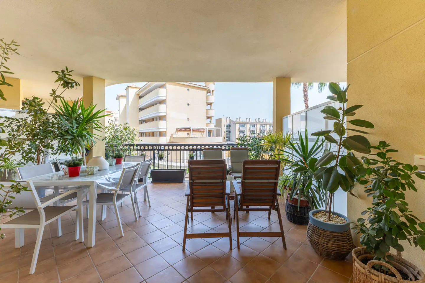 A sunlit balcony with brown tile flooring, a white table with chairs, wooden chairs, and potted plants.