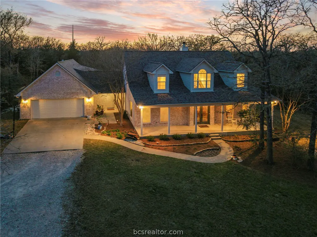 Exterior shot of a two-story brick house with a covered porch at dusk. The sky is pink and orange. A driveway leads to an attached garage.