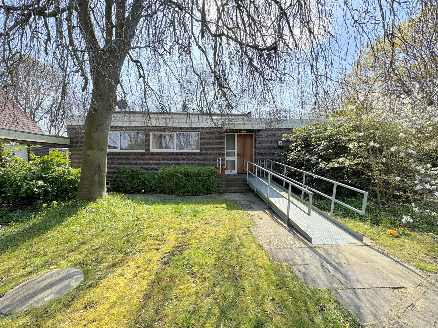 A single-story brick house with a ramp leading to the front door, surrounded by a green lawn and trees.
