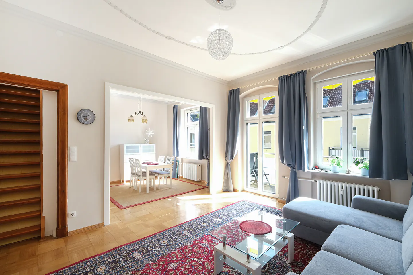 Bright living room with hardwood floors, a blue patterned rug, and a gray sofa. A dining room is visible through an archway.
