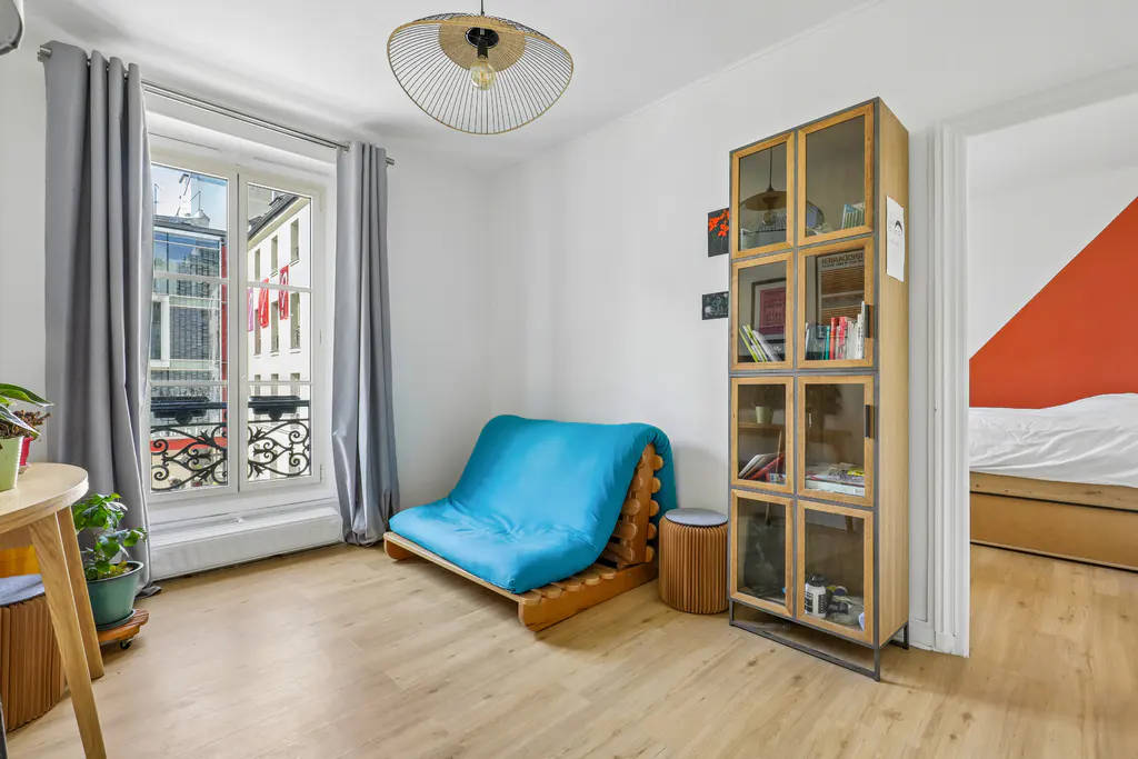 Bright living room with a blue futon, wooden bookshelf, and a window with gray curtains. A bedroom is visible through a doorway.
