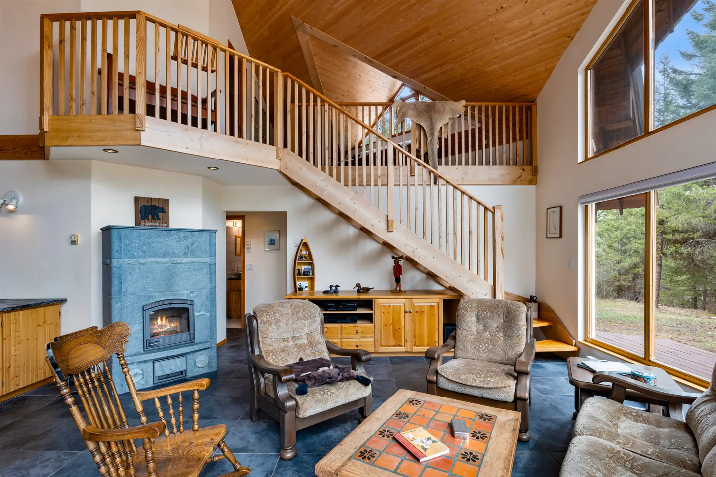 Living room with wood accents, a stone fireplace, and a staircase leading to a loft. Large windows overlook a wooded area.
