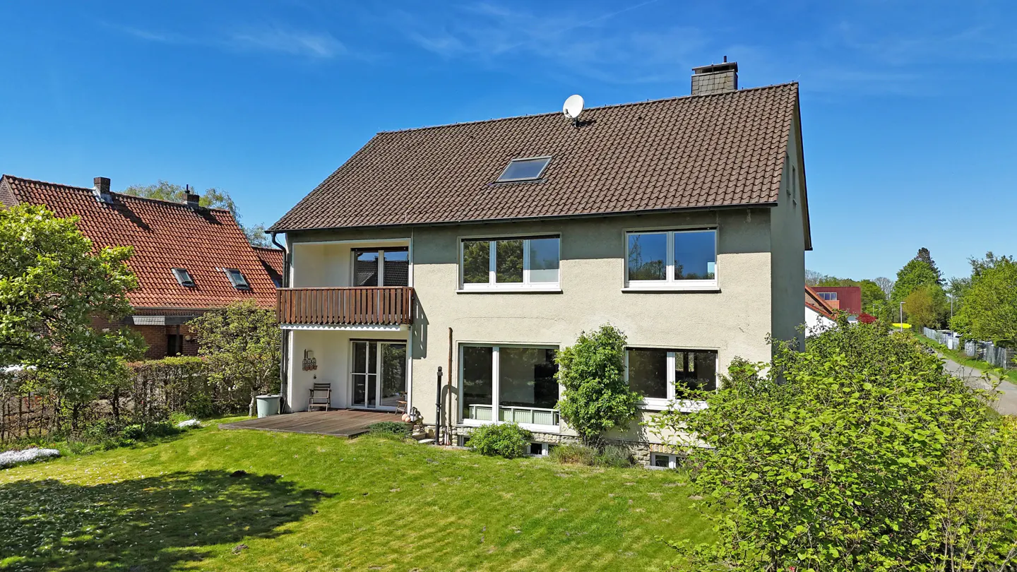 Two-story house with a brown roof, light-colored walls, a balcony, and a green lawn under a blue sky.