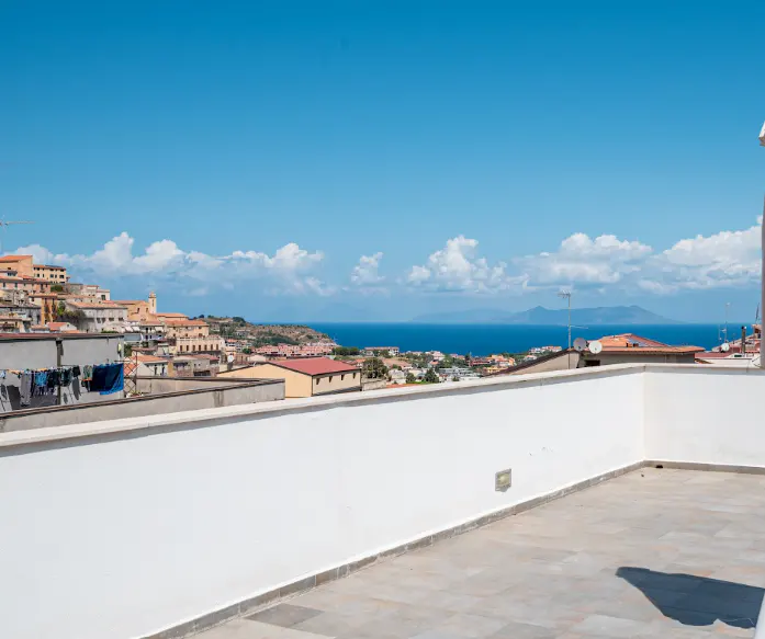 View from a rooftop terrace with a white wall overlooking a coastal town and the blue sea under a clear sky.