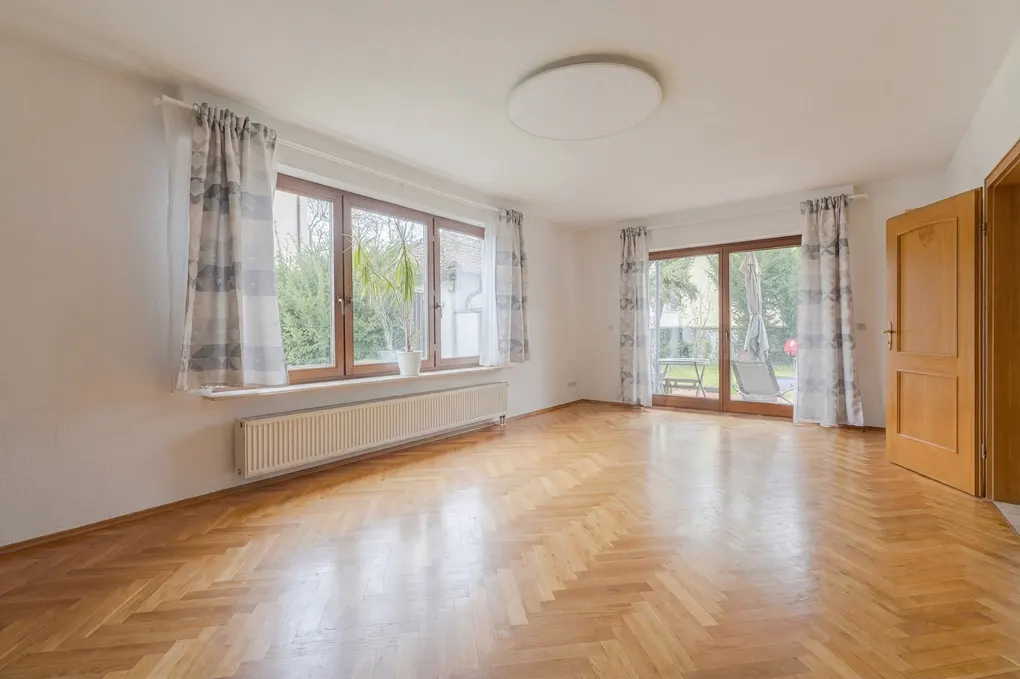 Bright, empty room with herringbone wood floors, white walls, and large windows with patterned curtains. A door leads outside to a patio.