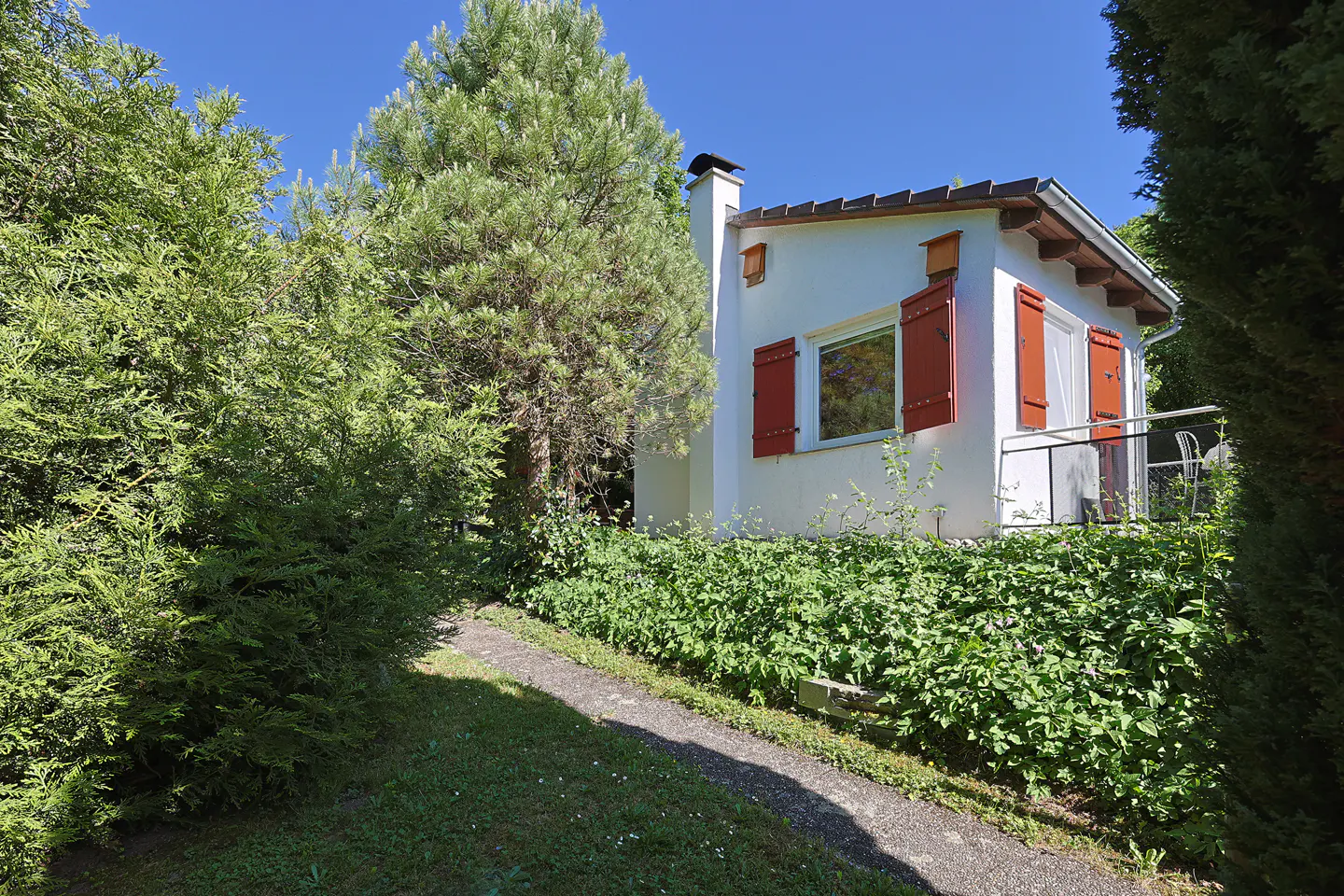 Small white cottage with red shutters and a stone path, surrounded by lush green trees and foliage under a clear blue sky.