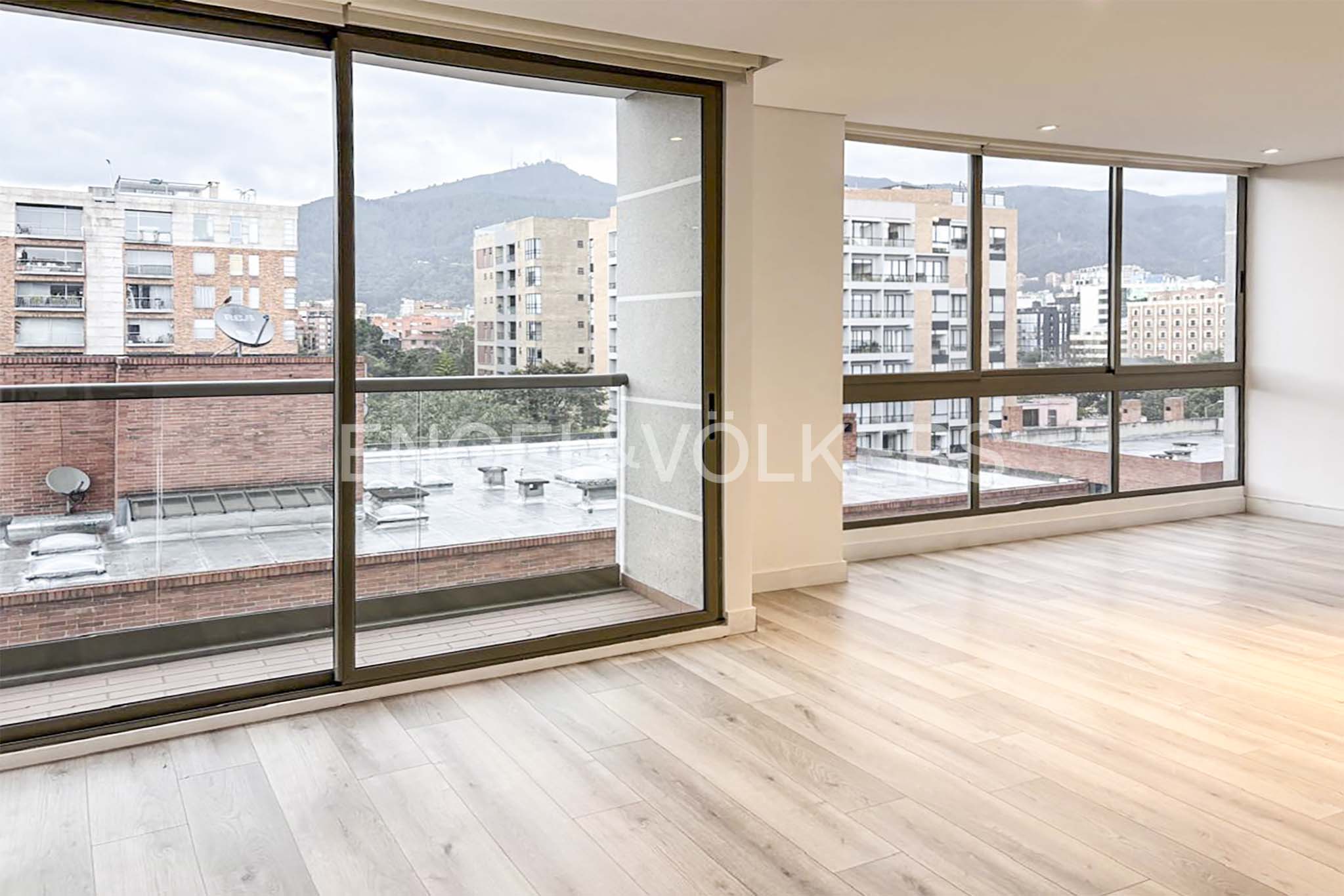 Bright, empty room with light wood floors and large windows. City buildings and a mountain are visible outside.