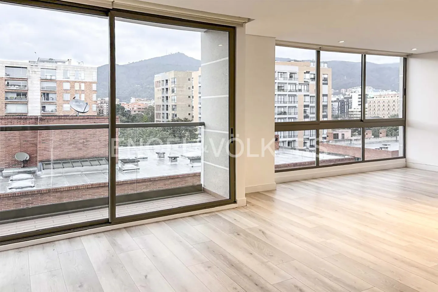 Bright, empty room with light wood floors and large windows. City buildings and a mountain are visible outside.