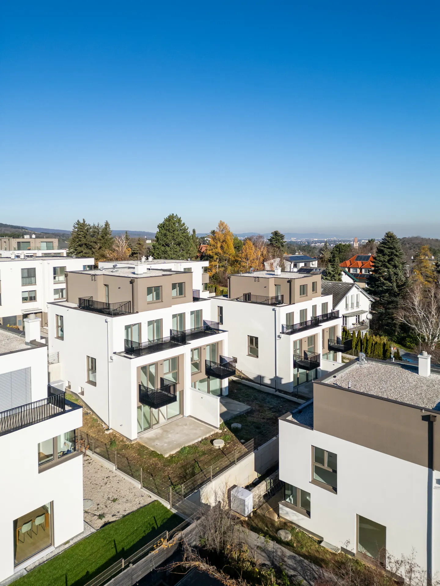Aerial view of modern white apartments with balconies, set against a clear blue sky and distant trees.