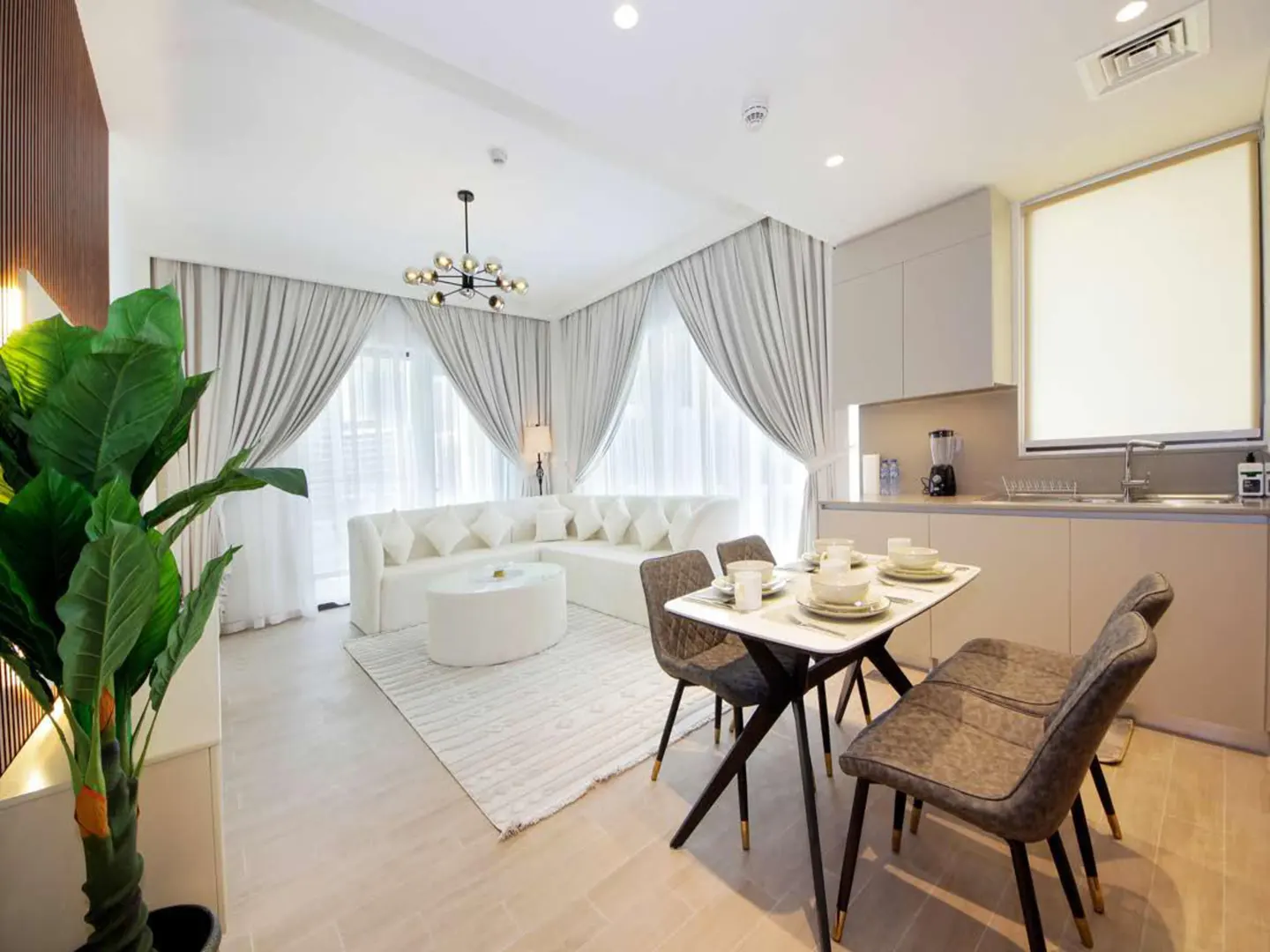 Bright, open-plan living space with white sofa, dining table set for four, and modern kitchen. Large windows with light gray curtains.