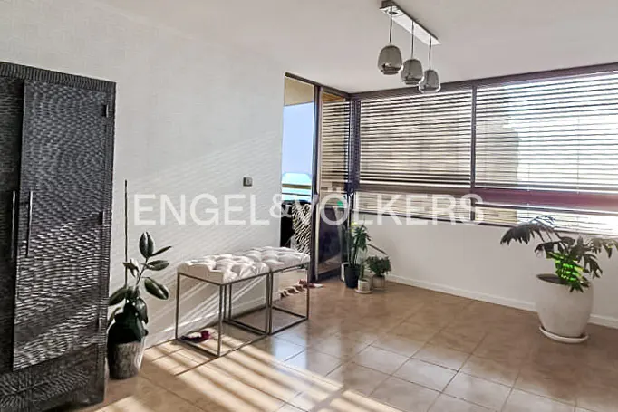 Bright room with tiled floors, two white ottomans, potted plants, and large windows with horizontal blinds.