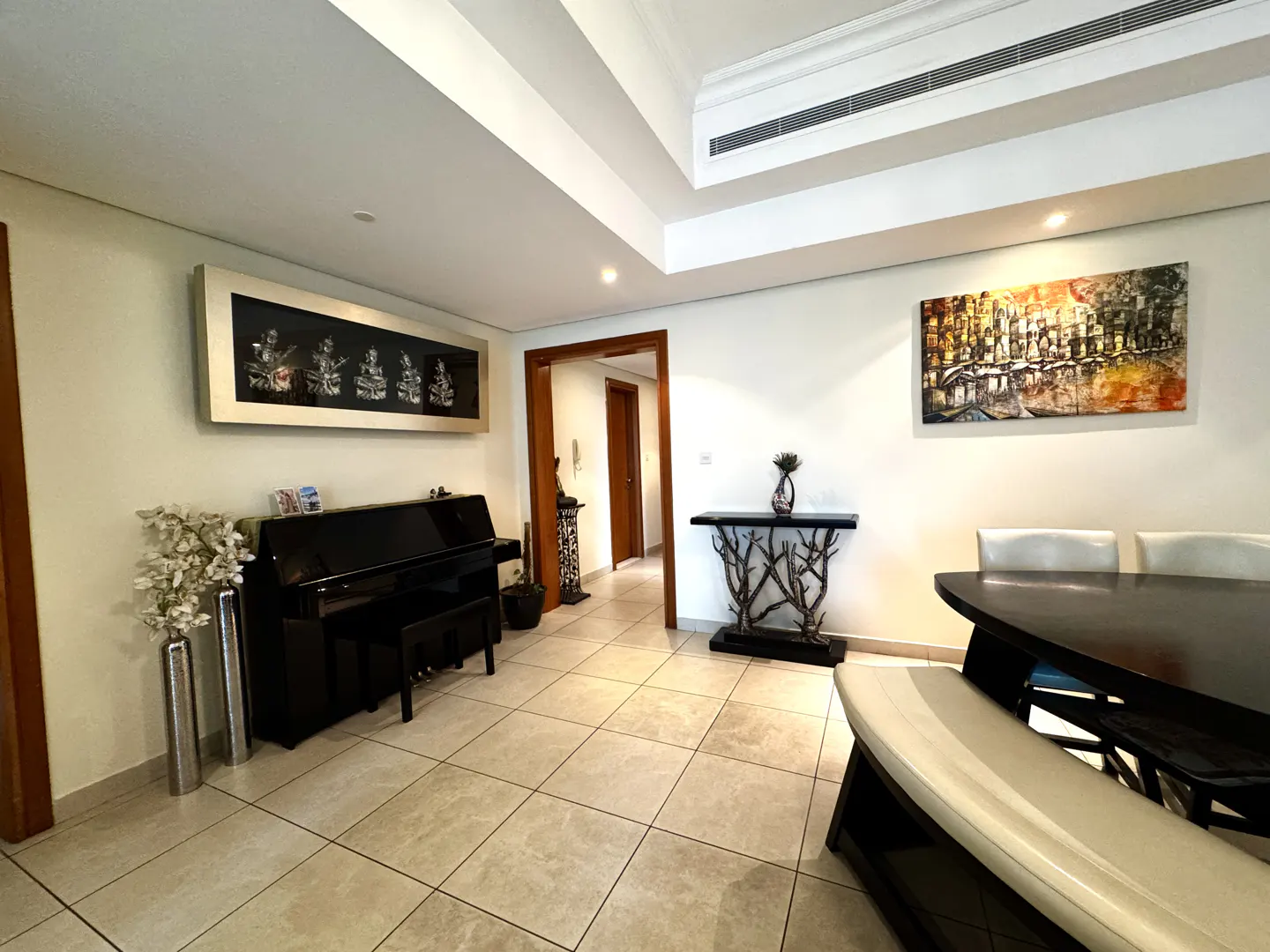A living room with a black piano, dining table, and artwork on the walls. The floor is tiled, and there is a doorway leading to another room.