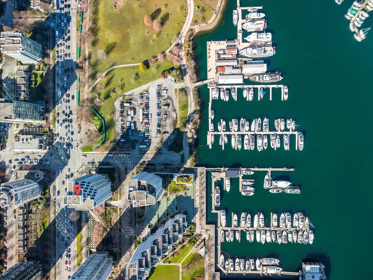 Aerial view of a marina with boats docked in green water, next to a city with buildings, a park, and a busy street.