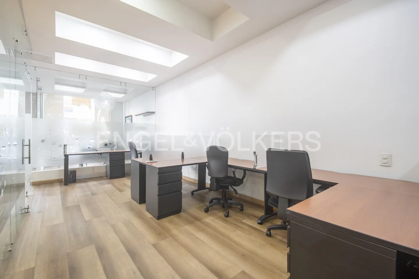 Bright office space with wood floors, white walls, and glass-enclosed area. Desks with black chairs are arranged throughout the room.