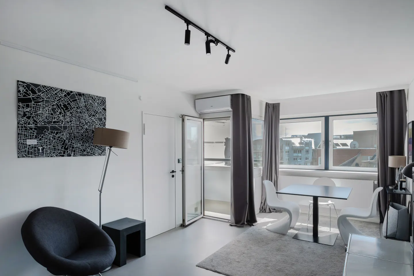 Bright, modern apartment interior with white walls, gray curtains, and a black and white city map art piece. A black chair and table are on the left. A table and chairs are on the right.