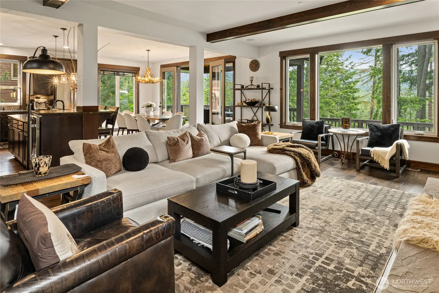 Open-concept living room with a large grey sectional, dark wood coffee table, leather armchair, and forest views.