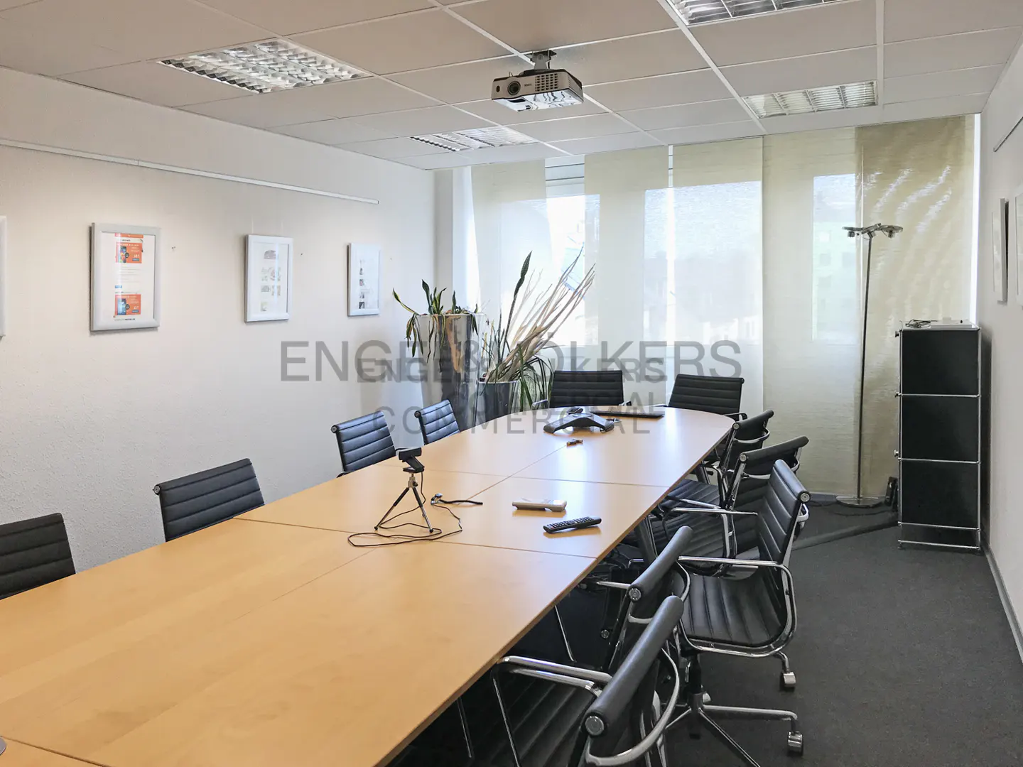 Conference room with a long wooden table, black chairs, projector, and plants. Natural light filters through the window blinds.