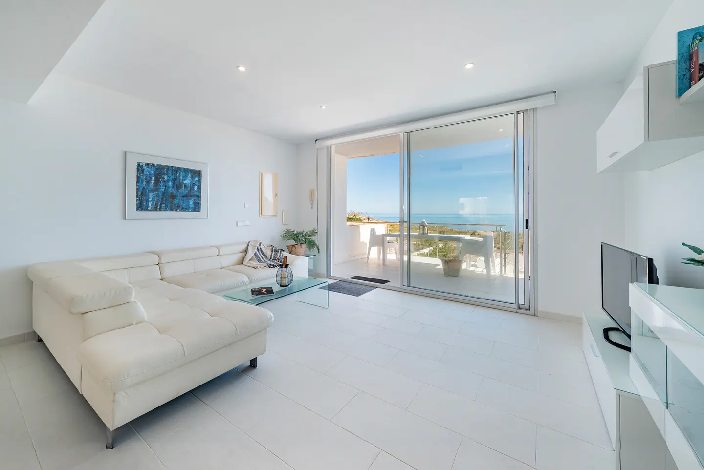 Bright, modern living room with white walls, tile floor, and a large sliding glass door to a balcony with ocean view. A white sectional sofa and glass coffee table are in the foreground.