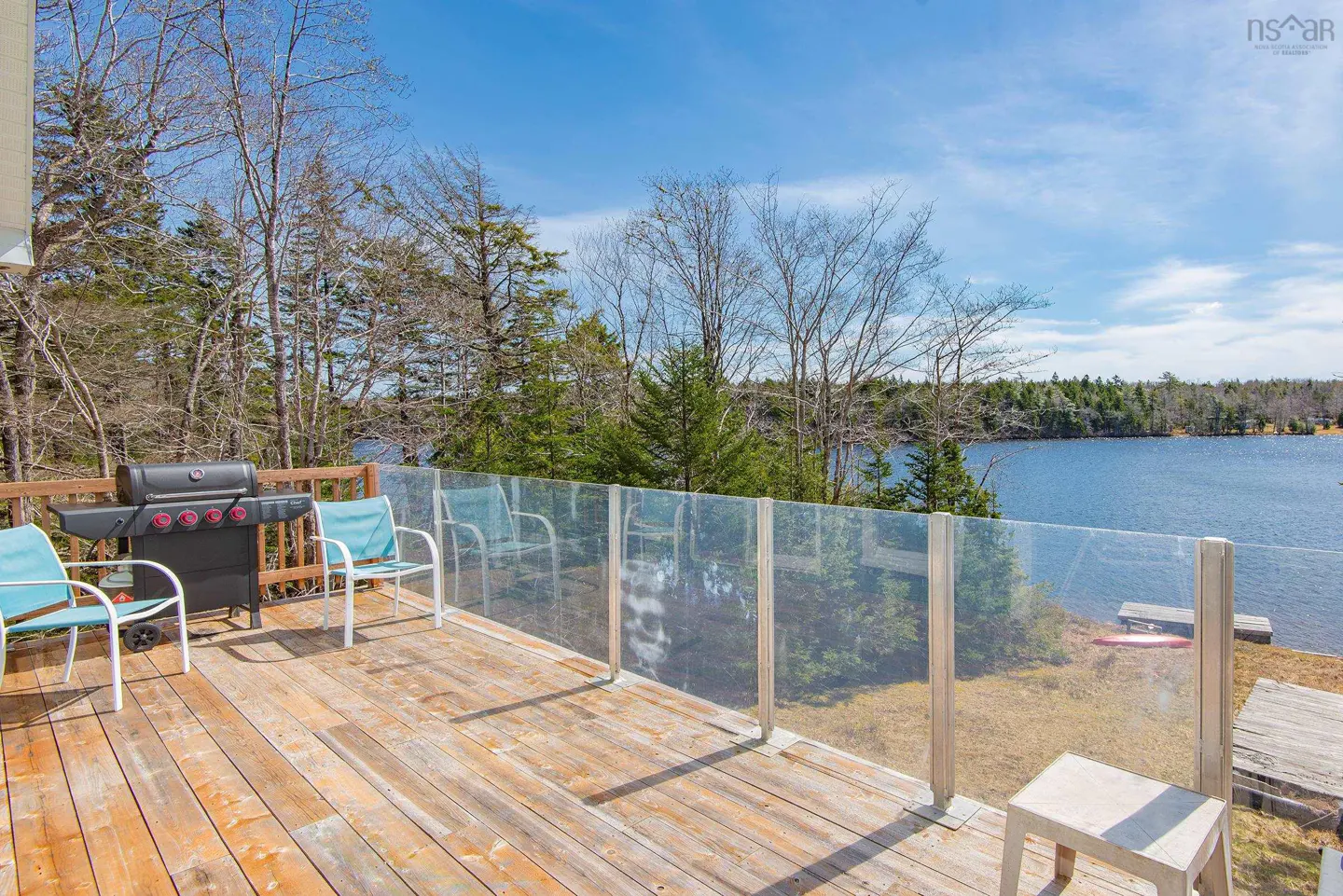A wooden deck with a grill, blue chairs, and a glass railing overlooks a lake surrounded by trees.
