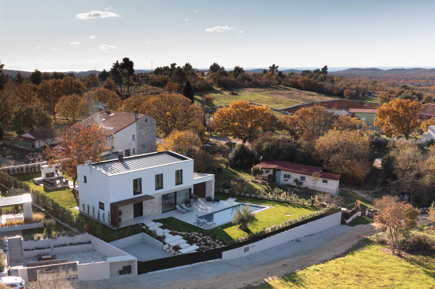 Aerial view of a modern, white two-story house with a pool and green lawn, surrounded by trees with autumn foliage.