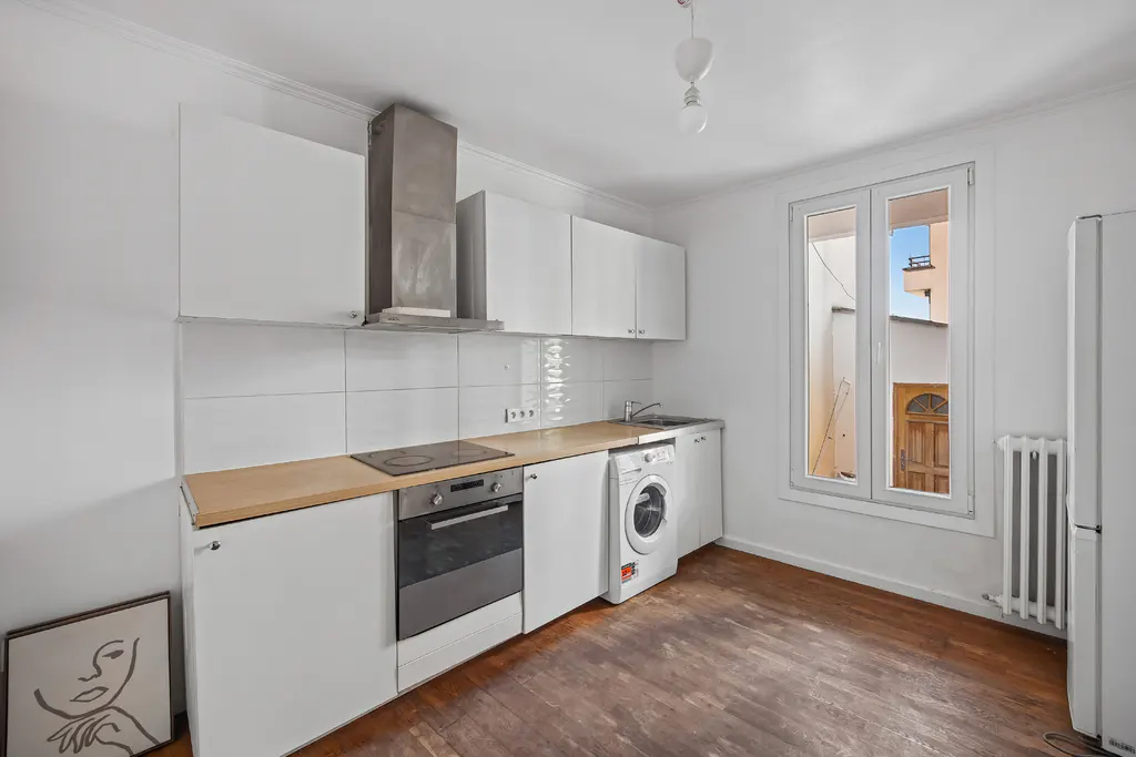 Bright kitchen with white cabinets, stainless steel hood, oven, and washing machine. A window overlooks a courtyard. Hardwood floor.