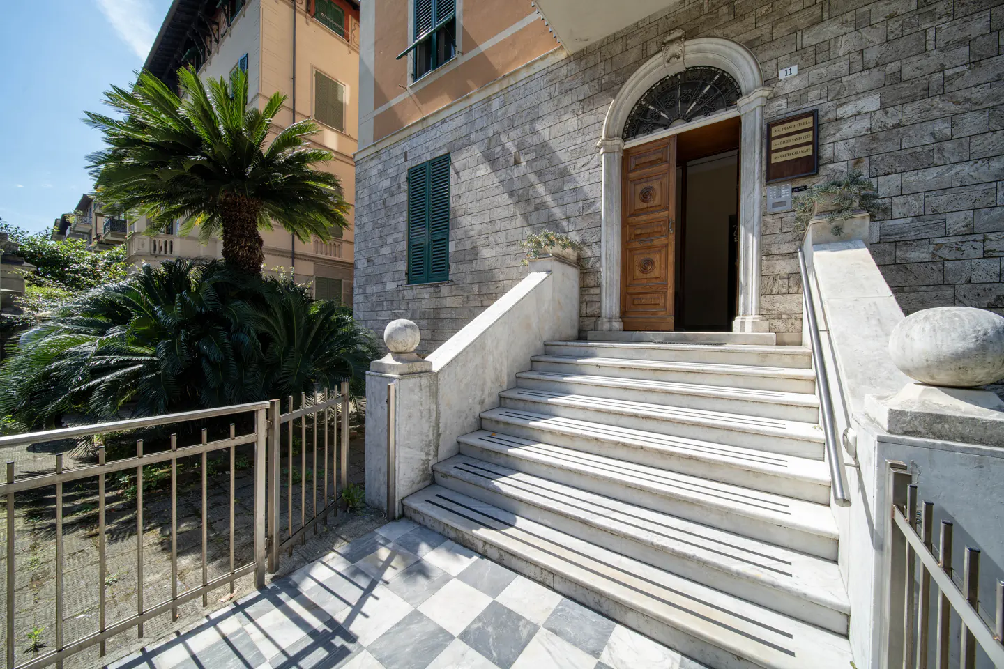 Exterior view of a stone building with marble steps leading to an open wooden door. A palm tree is on the left.